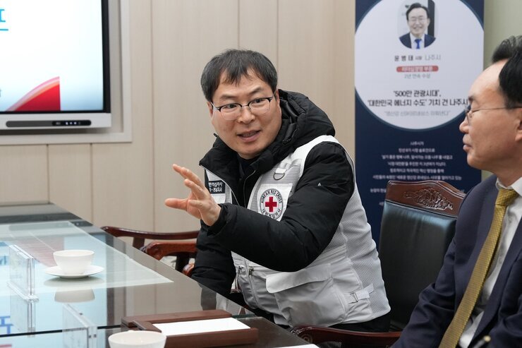 흰색 구호 조끼를 입은 중년 남성이 테이블 맞은편 사람에게 손을 사용해 설명하며 이야기하고 있는 모습