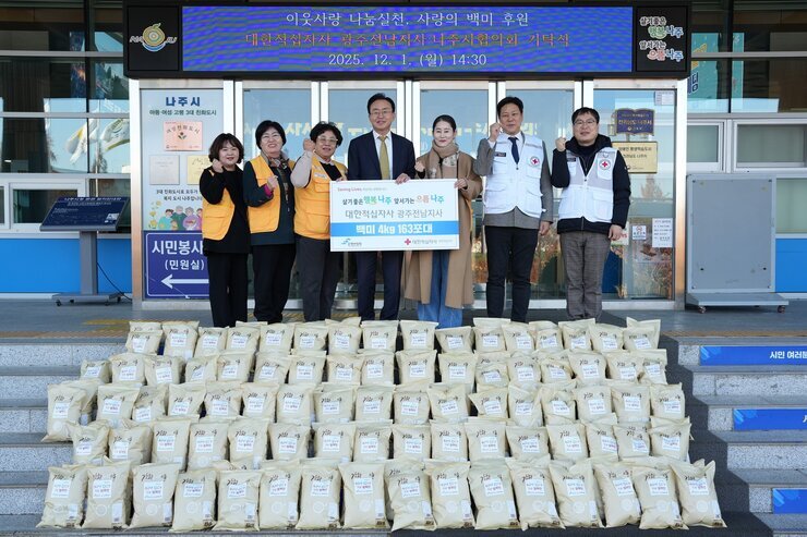 나주 시민청 앞에서 나주시장, 적십자 관계자 및 봉사자 7명이 '사랑의 쌀 163포(500만원 상당) 후원 기념' 플래카드를 들고 쌓여 있는 쌀 포대들 앞에서 포즈를 취하고 있는 전경. 상단 전광판에는 '이웃사랑 나눔실천, 사랑의 백미 후원' 행사가 '2025. 12. 1. (월) 14:30 ' 에 진행되었음이 표시되어 있다.