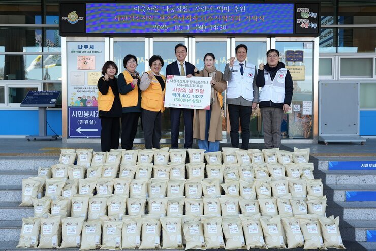 나주 시민청 앞에서 윤병태 나주시장(중앙), 적십자 관계자 및 봉사자 7명이 '사랑의 쌀 163포(500만원 상당) 후원 기념' 플래카드를 들고 쌓여 있는 쌀 포대들 앞에서 포즈를 취하고 있는 전경. 상단 전광판에는 '이웃사랑 나눔실천, 사랑의 백미 후원' 행사가 '2025. 12. 1. (월) 14:30 '에 진행되었음이 표시되어 있다.