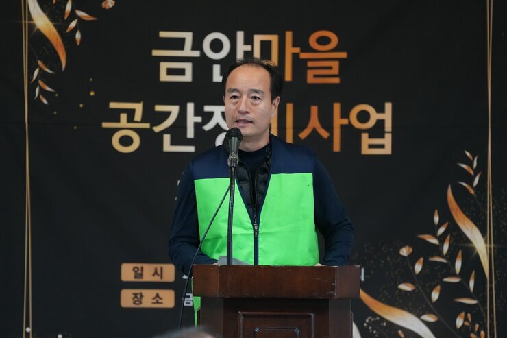  녹색 조끼를 입은 중년 남성이 단상에서 발언하는 모습. 배경에는 '금안마을 공간재생사업' 현수막이 보인다.