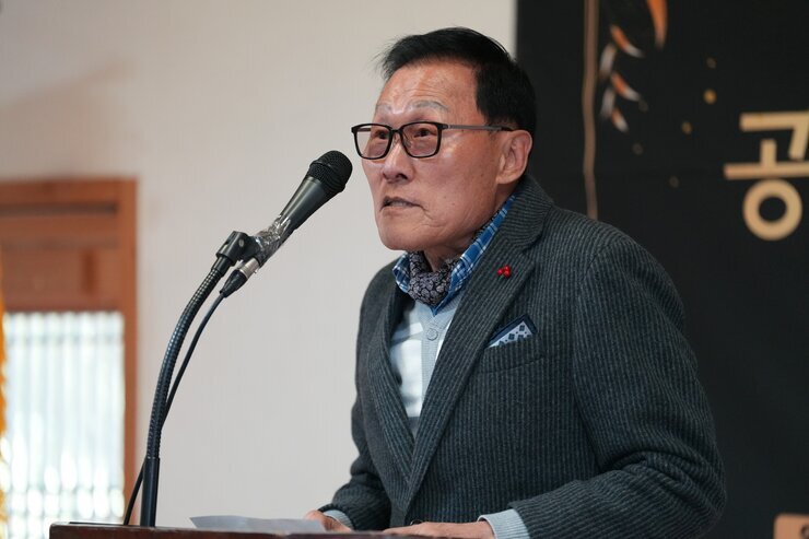  단상 앞에서 연설하는 노년 남성의 클로즈업 사진. 안경을 쓰고 재킷을 입었으며, 마이크 앞에서 발언하고 있다.