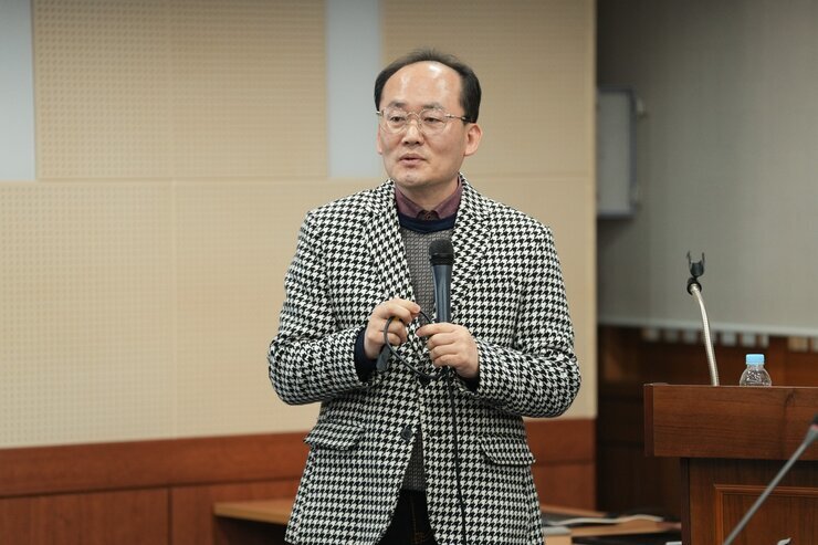  발표를 진행하는 중년 남성. 체크무늬 재킷을 입은 남성이 마이크를 잡고 이야기하고 있다.