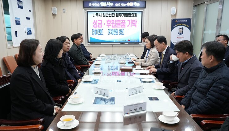  회의 또는 간담회 장면. 직사각형 테이블에 마주 앉은 나주시 관계자들과 기업 관계자들이 대화를 나누고 있다. 테이블 위에는 '조광호', '서영고', '나상채' 등의 명패가 놓여 있으며, 뒤쪽 화면에는 '나주시 일반산단 입주기업협의회 성금·후원물품 기탁 (300만원) (930만원 상당)'이라는 문구가 보인다.