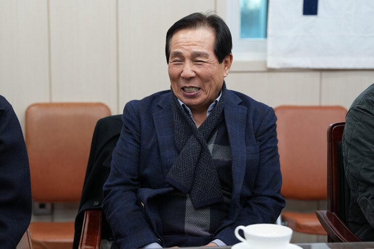  회의석상에 앉아 환하게 웃고 있는 노년 남성. 짙은 남색 재킷을 입고 체크무늬 목도리를 착용하고 있다.