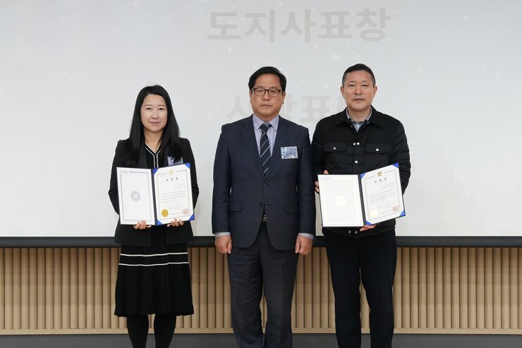  두 명의 수상자(여성 1명, 남성 1명)가 한 남성(중앙)과 함께 상장을 들고 기념 촬영하는 모습. 배경 스크린에 '도지사 표창'이 보임