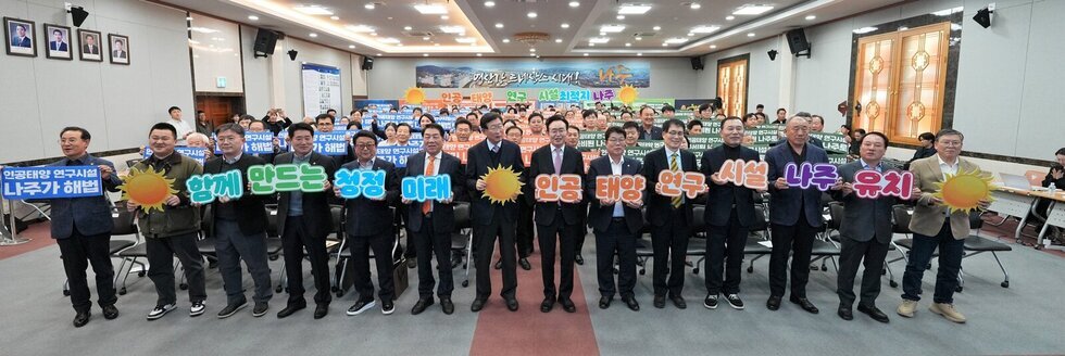 실내 회의장 같은 곳에서 정장을 입은 여러 남성들이 줄지어 서서 태양 모양 피켓과 플래카드를 들고 있는 모습. 플래카드에는 '인공태양 연구시설 나주가 해법', '함께 만드는 성장 미래', '인공태양 연구시설 나주 유치' 등의 문구가 적혀 있다.