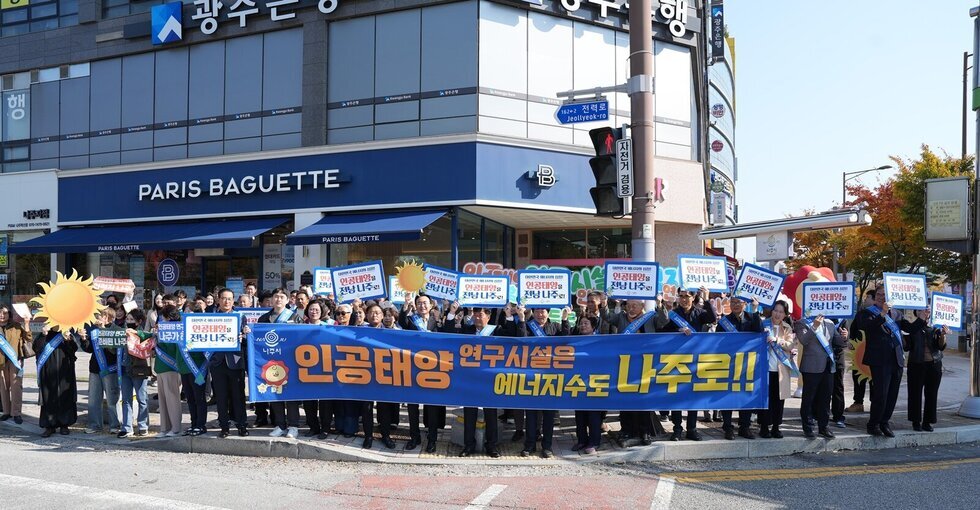 길거리에서 사람들이 'PARIS BAGUETTE' 매장 앞을 배경으로 단체로 서서 인공태양 연구시설 유치를 촉구하는 피켓과 긴 플래카드를 들고 있는 모습. 긴 파란색 플래카드에는 '인공태양 연구시설은 에너지수도 나주로!!' 라고 크게 적혀 있다. 사람들은 태양 모양 피켓과 '인공태양을 전남 나주로' 등의 문구가 적힌 피켓을 들고 있다.