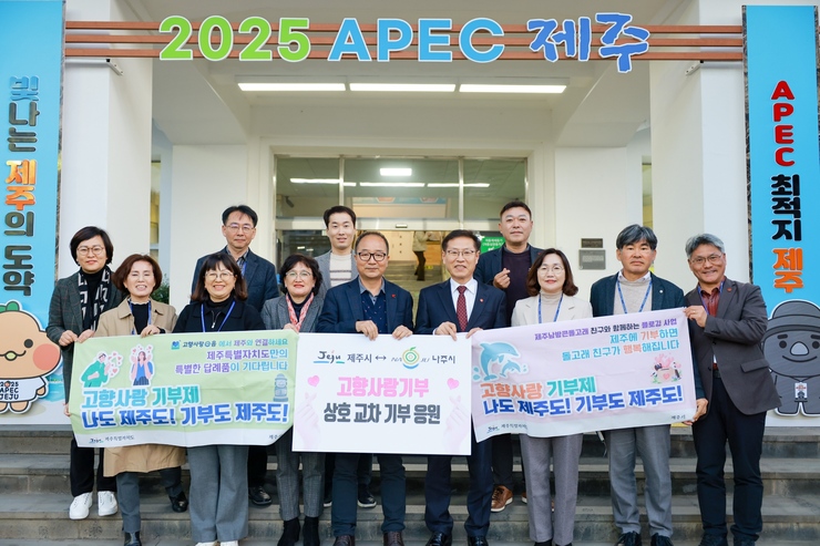 건물 상단에 2025 APEC 제주 라고 적혀 있고 고향사랑 기부제 나도 제주도! 기부도 제주도! 라고 적힌 현수막을 들고 기념촬영하고 있다.