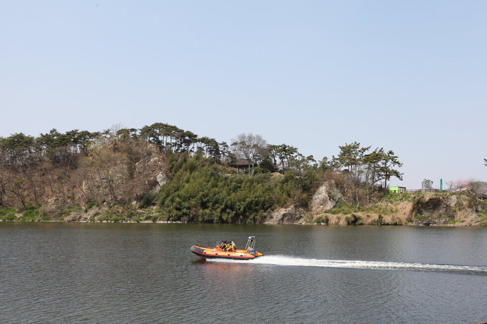 영산강 위에 보트가 보이고 그 뒤로 수풀이 우거진 돌산이 보이며 그 안에 석관정이 있음.
