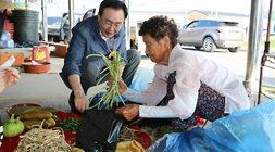 추석 명절맞이 나주목사고을시장 장보기 행사 나주시장이 쪼그려 앉아 검정비닐을 벌리고 있고 판매하는 여성 어르신이 나물을 담고 있는 모습
