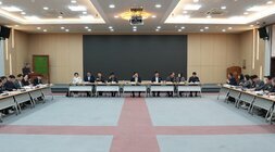 넓은 회의장에서 'ㄷ'자 형태로 테이블에 둘러앉아 회의하는 많은 사람들의 모습. 중앙에는 주요 관계자들이, 양 옆 긴 테이블에는 여러 참석자들이 앉아있다. 뒤쪽 벽에는 검은색 스크린과 액자들이 걸려있다.