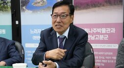 정장을 입고 테이블에 앉아 있는 중년 남성. 흰 셔츠와 짙은 색 재킷을 착용하고 있으며, 안경을 쓰고 정면을 바라보고 있다.