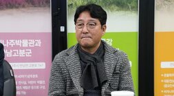  테이블에 앉아 있는 중년 남성. 목도리와 체크재킷을 착용하고 있으며, 안경을 쓰고 정면을 바라보고 있다.