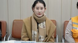 카멜색 스카프를 두른 여성이 정면을 응시하며 회의 테이블에 앉아 있는 클로즈업 사진