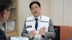 흰색 구호 조끼를 입은 중년 남성이 테이블 맞은편 사람에게 손을 사용해 설명하며 이야기하고 있는 모습