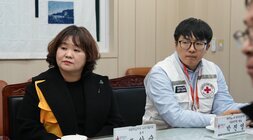 회의 테이블에 나란히 앉아 있는 두 참석자. 왼쪽의 여성은 검은색 외투를 입었고, 오른쪽의 남성은 흰색 조끼를 입고 안경을 쓰고 있다.