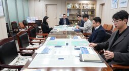 회의실 테이블에 앉아 있는 여러 명의 참석자들. 테이블 위에는 지도나 보고서가 펼쳐져 있다.