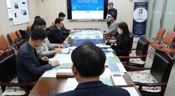 넓은 회의실에서 여러 명이 테이블을 둘러싸고 앉아 회의를 하고 있는 전경. 정면의 스크린에는 '중간보고' 관련 제목이 보이고, 한 남성이 스크린 옆에서 자료를 들고 서 있다.