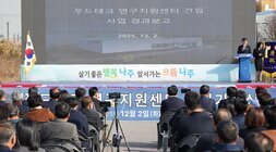 푸드테크 연구지원센터 건립공사 사업 경과보고 행사 전경. 야외 무대 위 스크린에 '푸드테크 연구지원센터 건립 사업 경과보고'와 센터 조감도가 표시되어 있다. 태극기 옆 단상에서 남녀 두 사람이 서 있으며, 앞줄에는 많은 참석자들이 의자에 앉아 경청하고 있다.