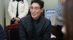 회의석상에 앉아 웃고 있는 중년 남성. 검은색 집업 재킷을 입고 안경을 쓰고 있다.