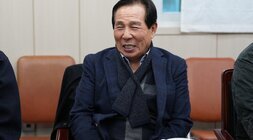 회의석상에 앉아 환하게 웃고 있는 노년 남성. 짙은 남색 재킷을 입고 체크무늬 목도리를 착용하고 있다.