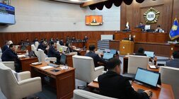 시의회 본회의장 전경. 의원들이 책상에 앉아 있고, 중앙 단상에서 발언하는 사람이 있으며, 벽면 스크린에는 '의회' 글자와 회의 내용이 보임