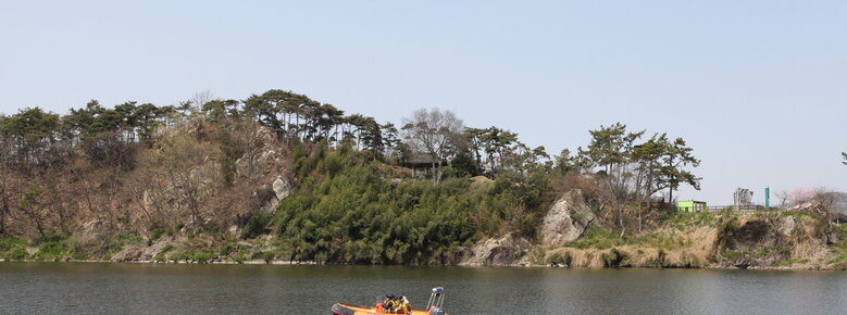 영산강 위에 보트가 보이고 그 뒤로 수풀이 우거진 돌산이 보이며 그 안에 석관정이 있음.